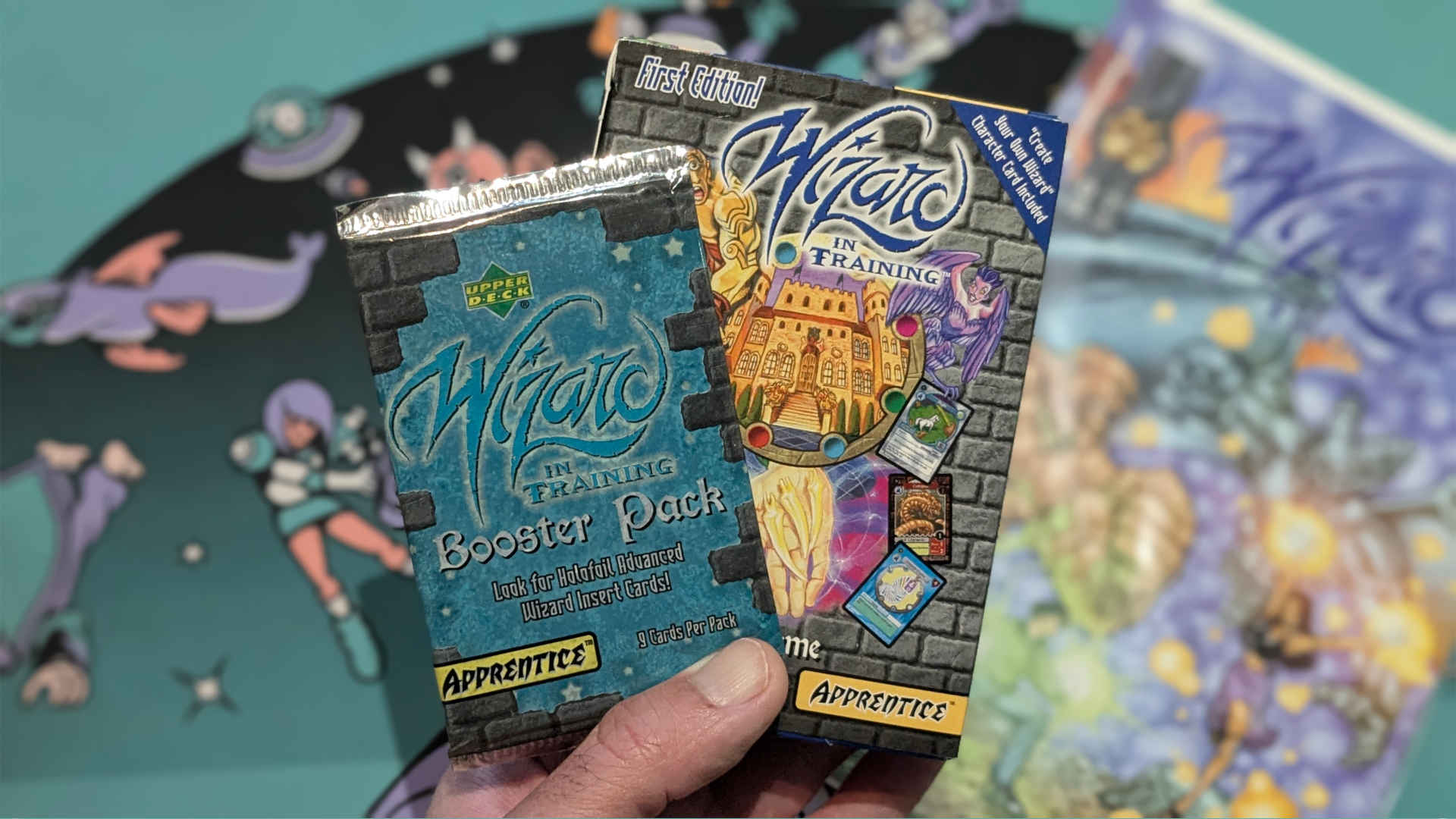 Wizard in Training TCG booster pack and starter deck, held in a hand above a playmat