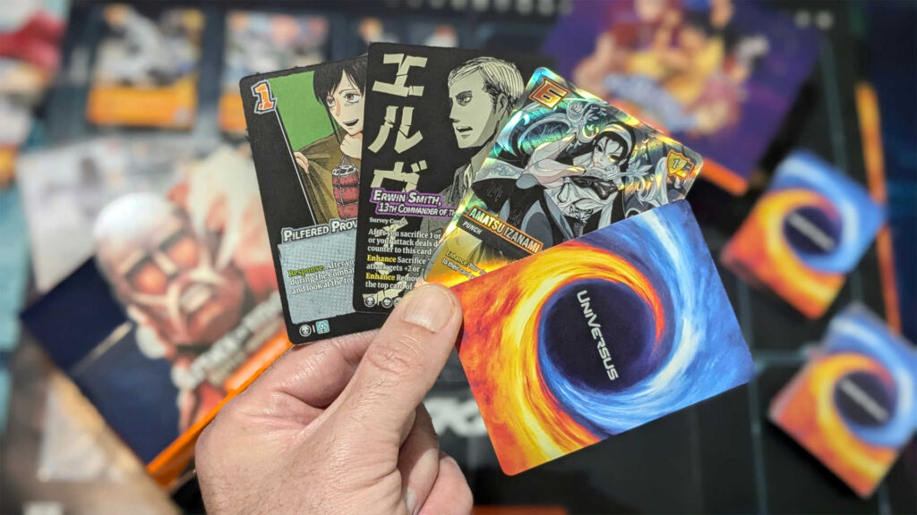 A hand of UniVersus cards fanned out, with cards and booster boxes out of focus in the background